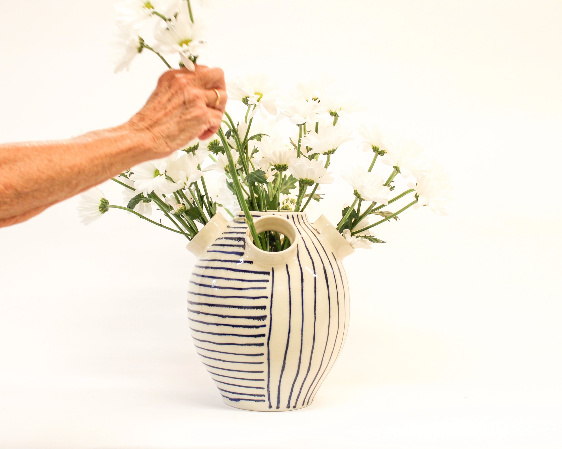 Person arranging white flowers into a handmade striped ceramic vase on a white background from Mima Ceramics by Michelle Organ.