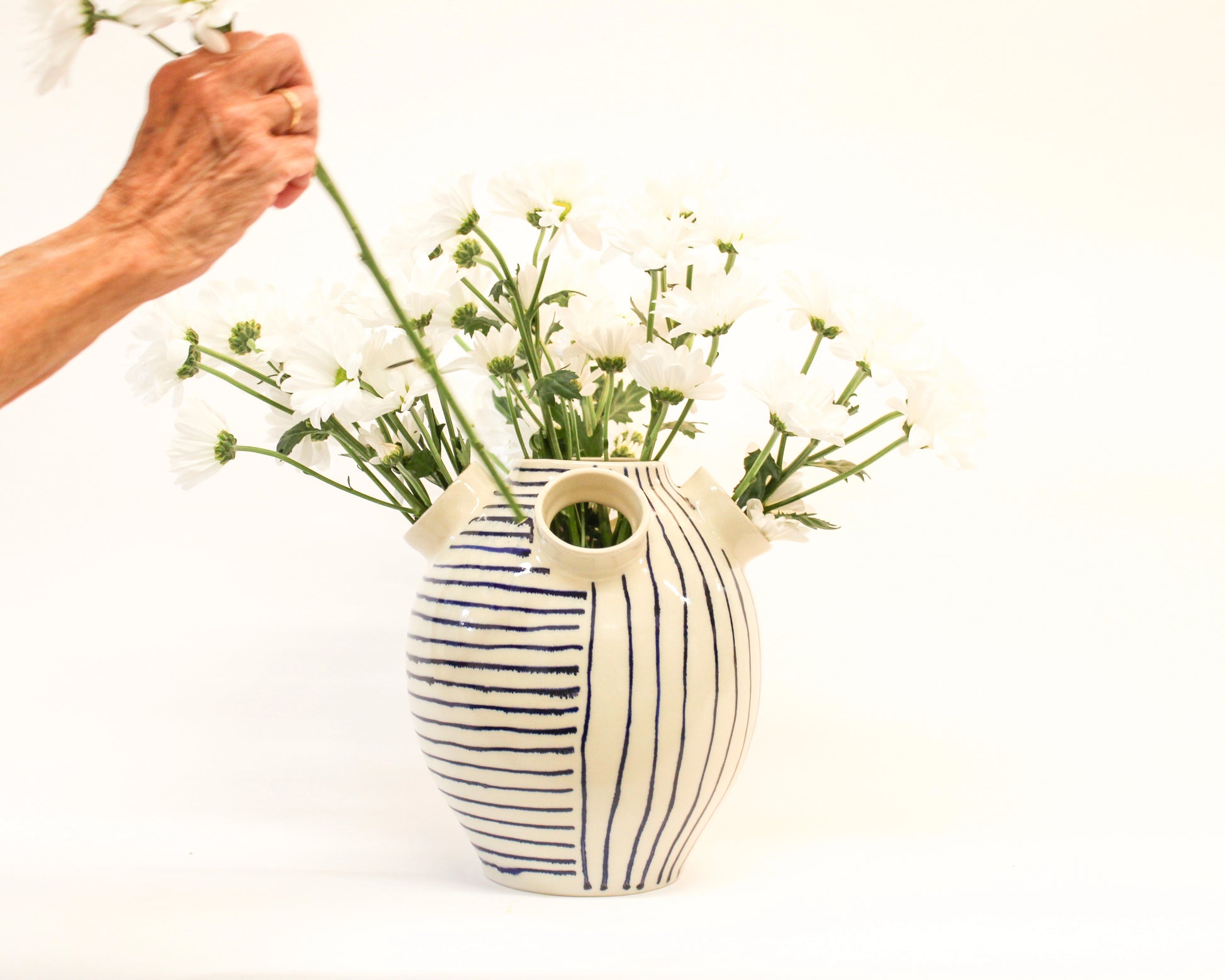 Hand arranging flowers into a handmade striped ceramic vase on a white background from Mima Ceramics by Michelle Organ. 