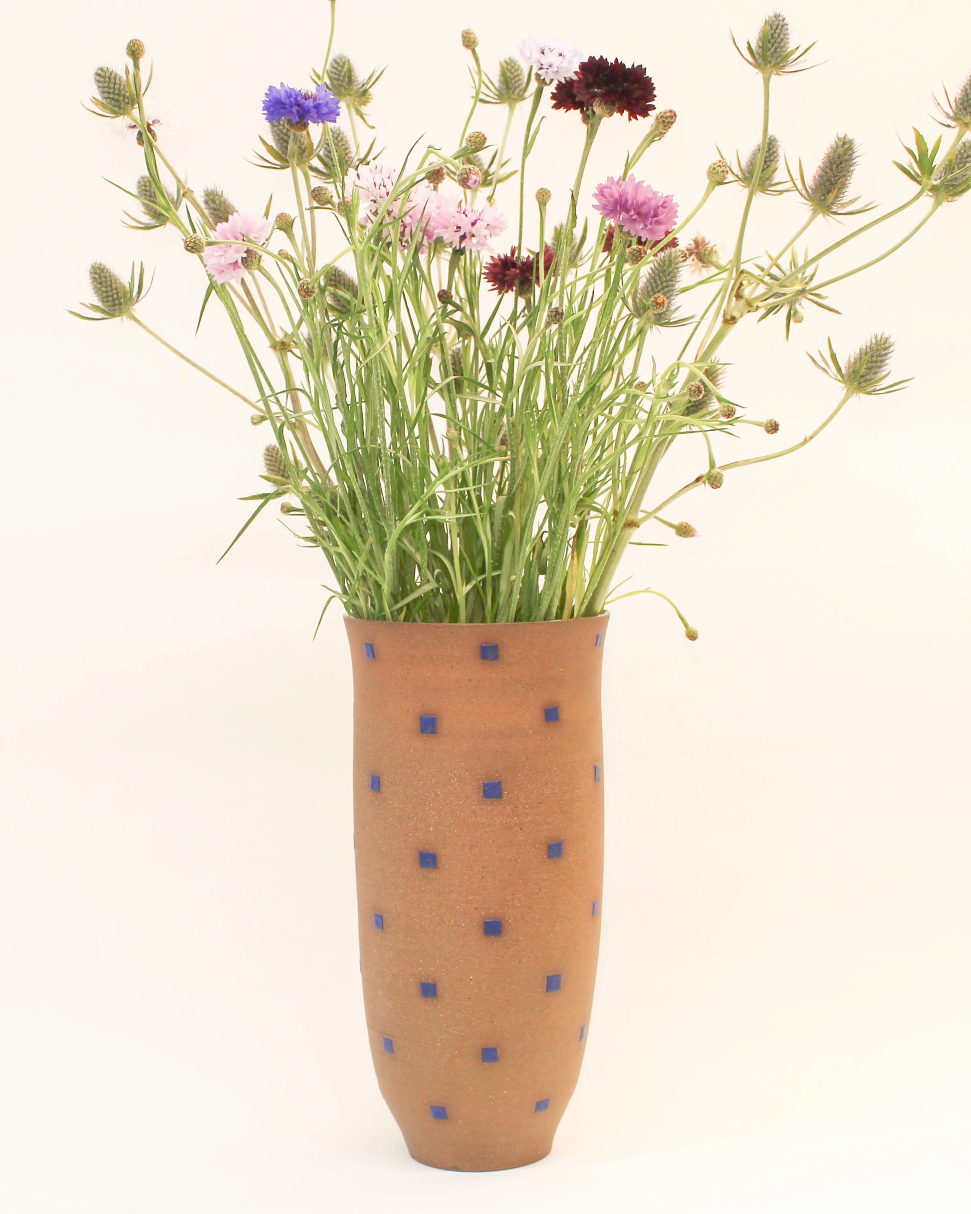Handmade Tall Red vase with a blue square pattern containing a bouquet of wildflowers on a white background by Mima Ceramics by Michelle Organ.